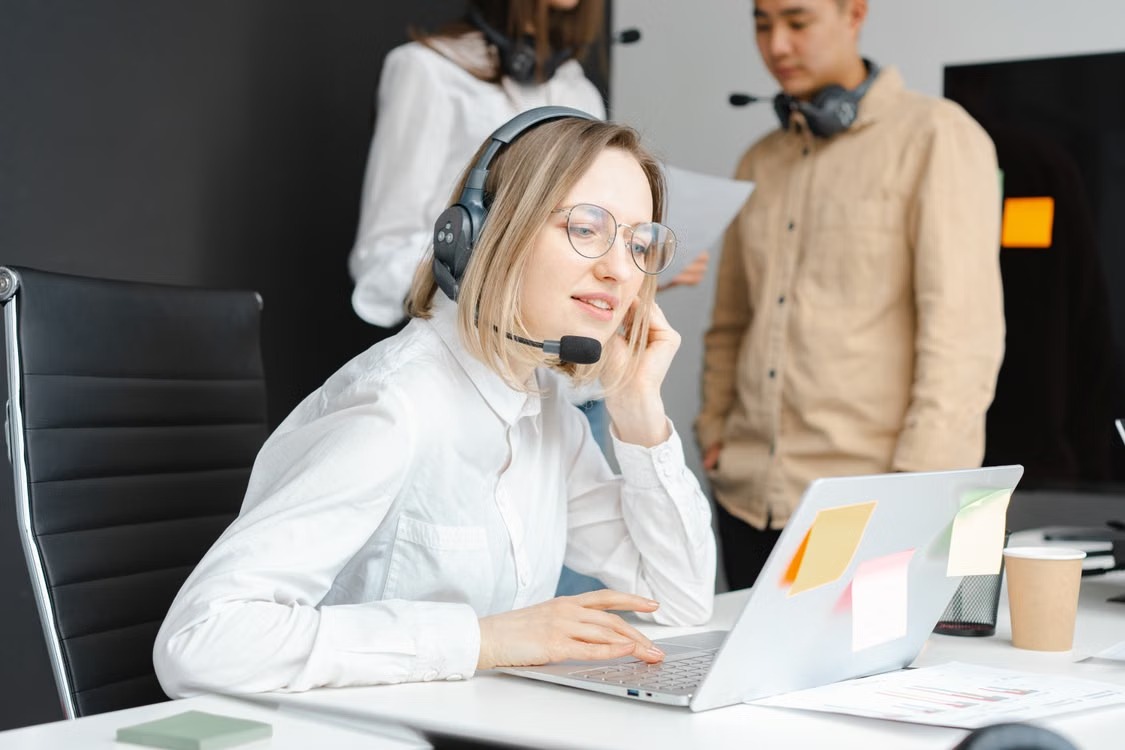 A support professional wearing a headset and working at a laptop with colleagues nearby.